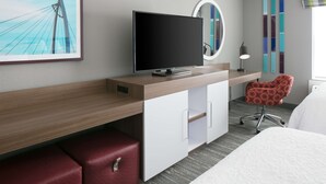 Une chambre d’hôtel moderne avec un bureau en bois, une télévision, un pouf en cuir rouge, un miroir rond, une chaise rouge et un lit.