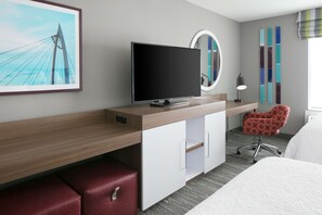 A modern hotel room with a wooden desk, a television, a red leather ottoman, a round mirror, a red chair, and a bed. - Hampton Inn Wichita Northwest (Wichita)