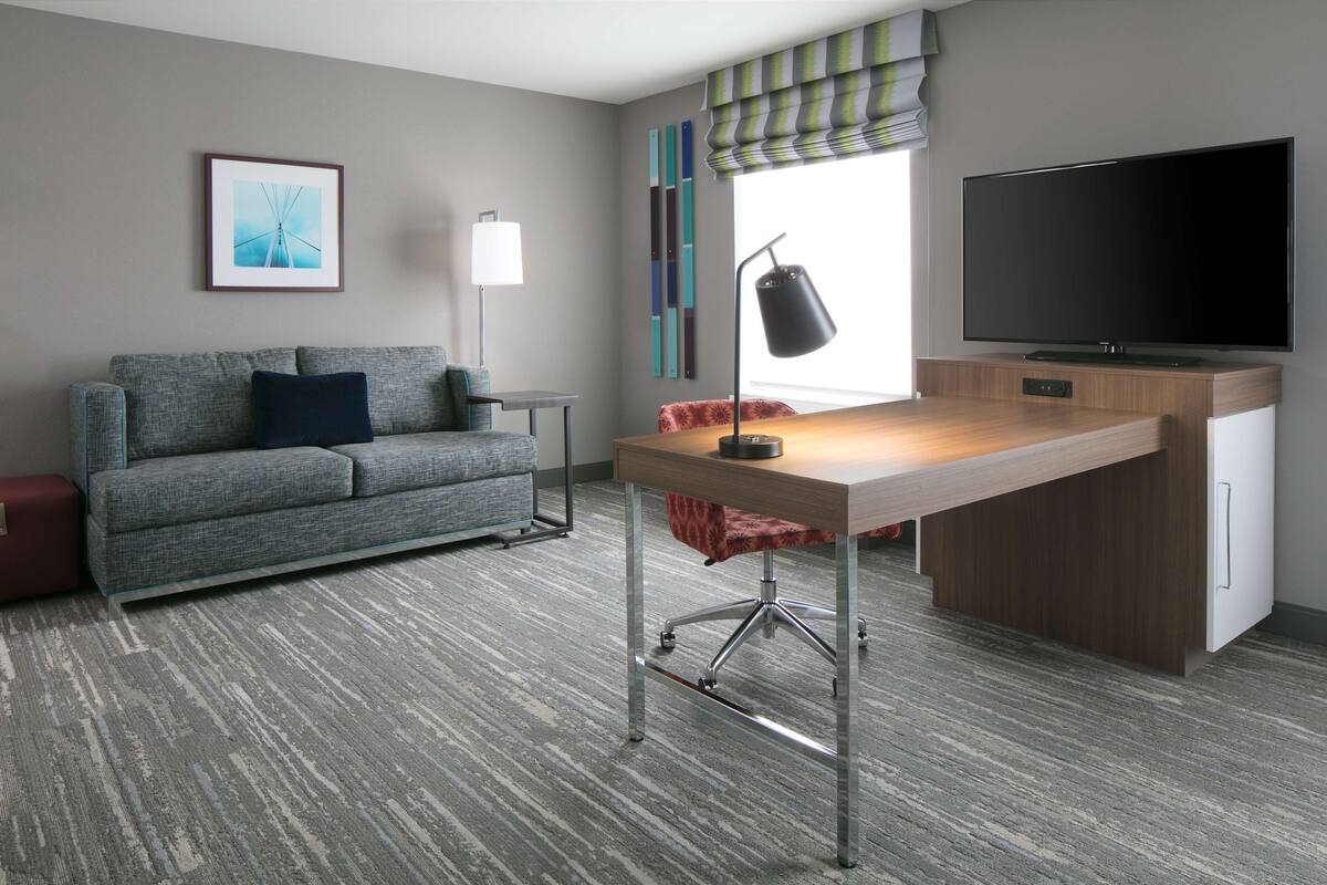 Une chambre d’hôtel moderne avec un canapé gris, un bureau en bois, un téléviseur à écran plat, une lampe sur pied et une fenêtre avec des rideaux à rayures.