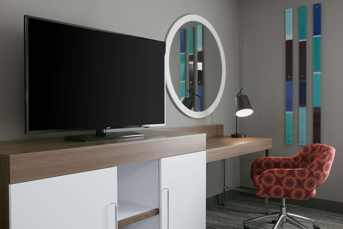 Une chambre d’hôtel moderne dotée d’une télévision à écran plat, d’un miroir rond, d’un bureau avec une lampe et d’une chaise à motifs.