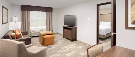 A hotel room with a sofa, ottoman, TV, and a bed with a headboard.