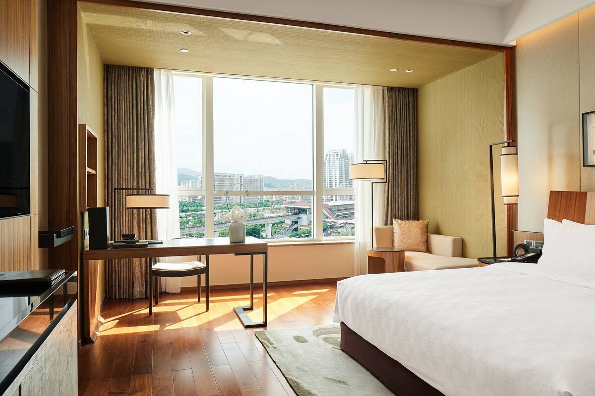 Une chambre d’hôtel avec un grand lit, un bureau avec une chaise, une télévision et une vue sur la ville grâce à de grandes fenêtres.