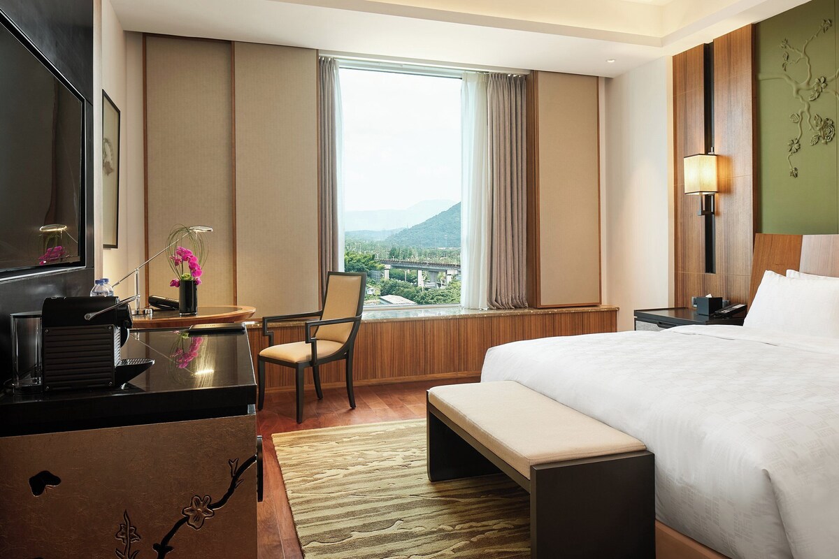 A hotel room with a large bed, a desk with a chair, a lamp, and a view of a bridge and mountains through the window.