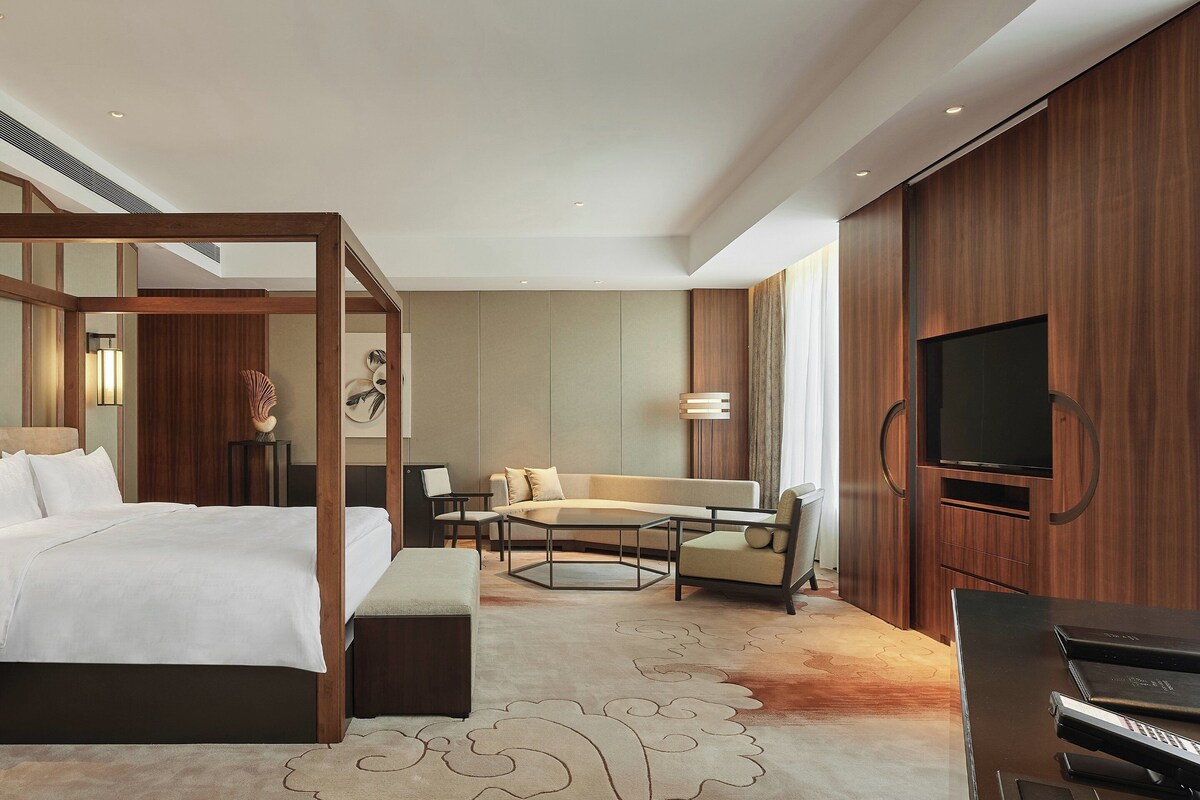 A modern hotel room with a four-poster bed, a sofa, a TV, and a desk with a chair.