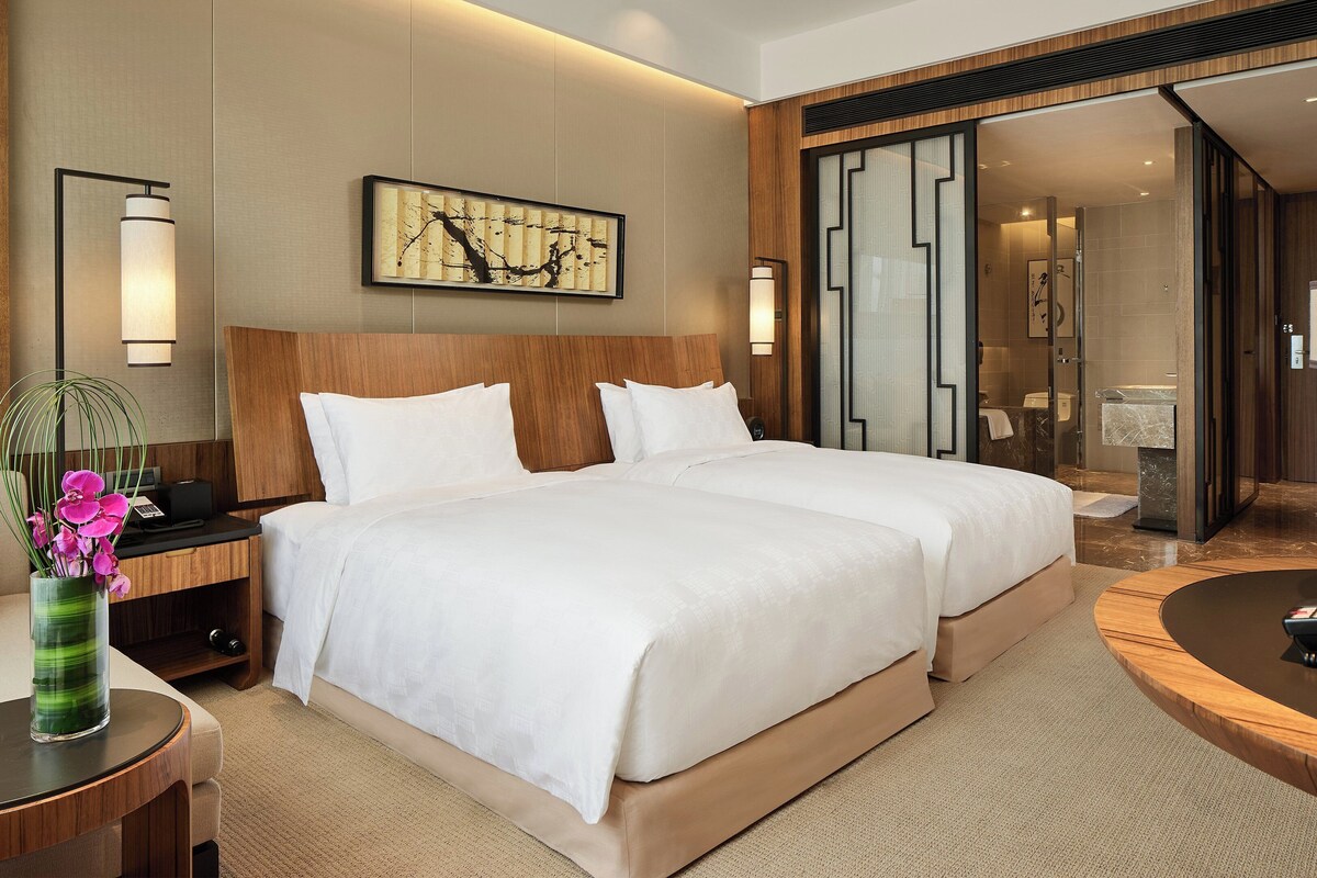 A hotel room with a large bed, a desk with a lamp, a vase with flowers, and a painting on the wall.