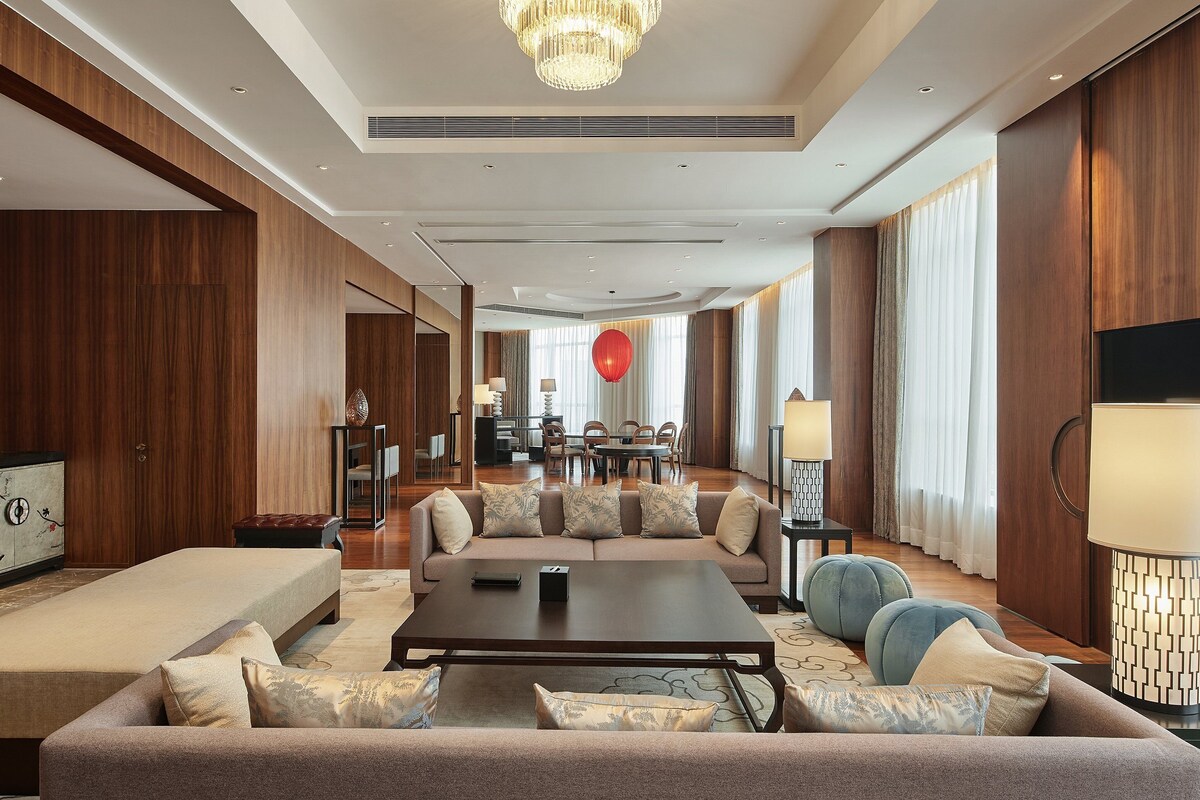 A spacious living area with a sofa set, a coffee table, and a TV. There is a dining area in the background with a red spherical decoration.