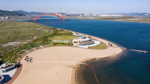 An aerial view of a coastal area with a large sandy beach, a modern building with a circular design, and a red bridge in the background. - HUALUXE Ningbo Harbor City by IHG (Ningbo)
