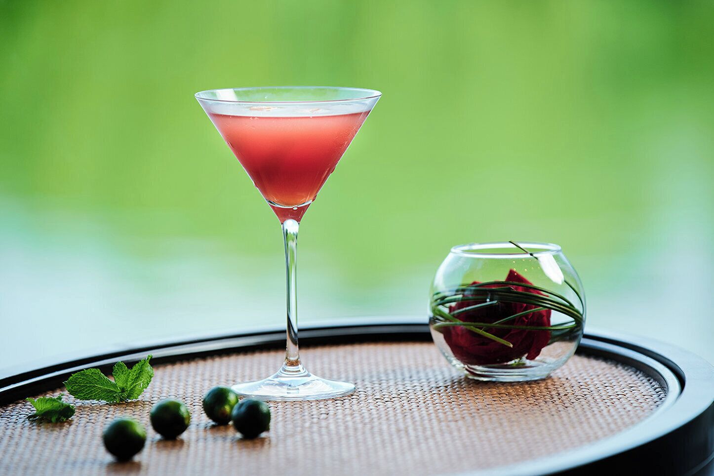 A martini glass with a red drink on a table with a woven placemat, a small glass with a rose, and some green leaves.