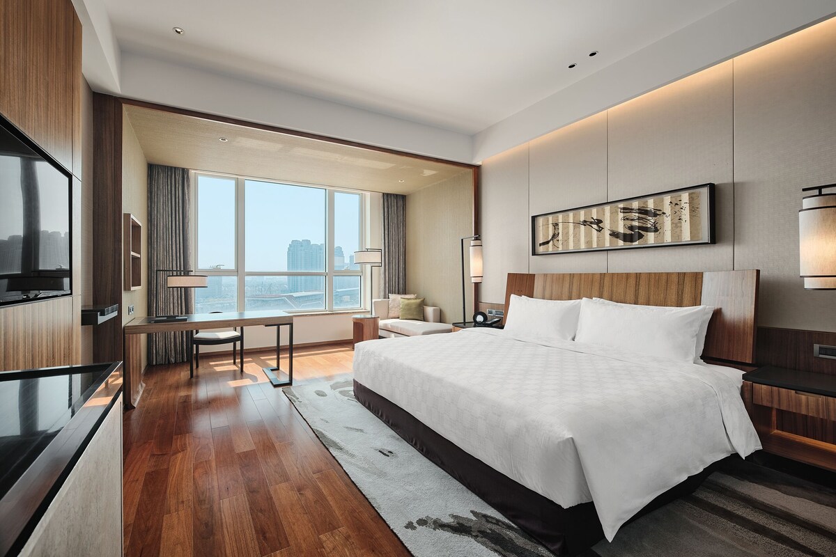 A modern hotel room with a large bed, a desk, and a view of the cityscape.
