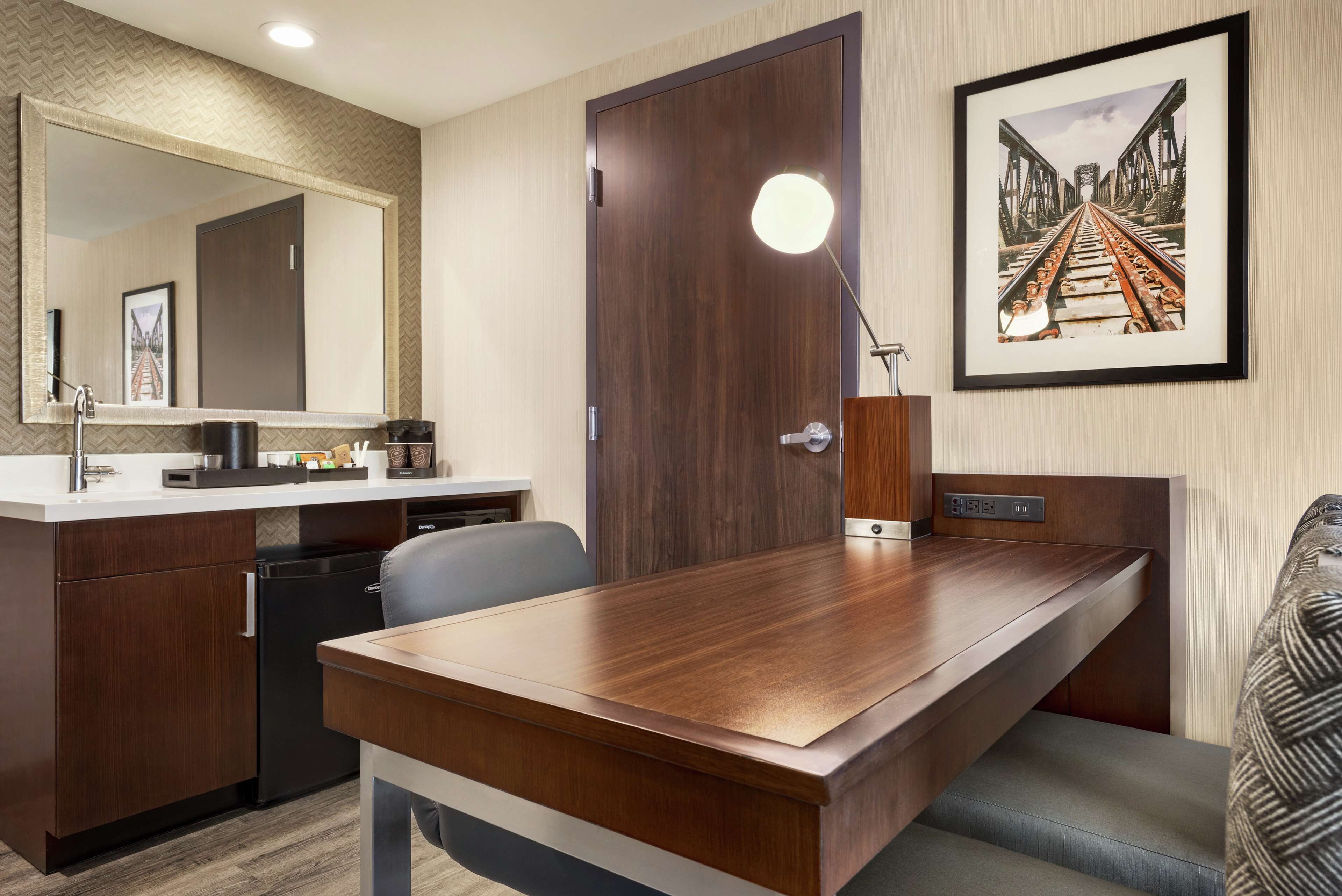 A modern hotel room with a wooden desk, a large mirror, a framed picture on the wall, and a built-in TV.