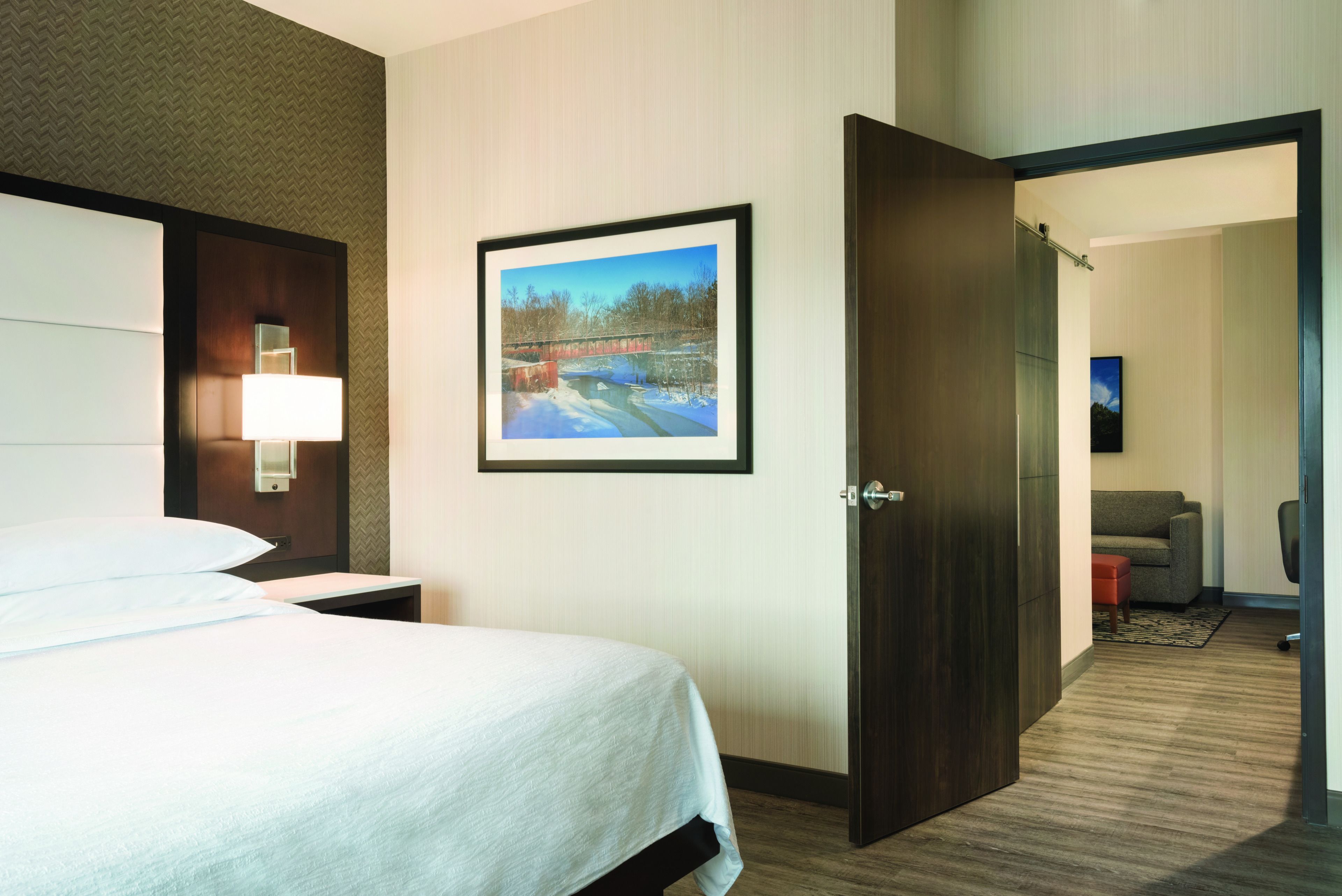 Una habitación de hotel con cama, mesita de noche, un cuadro en la pared y una puerta que da a otra habitación.