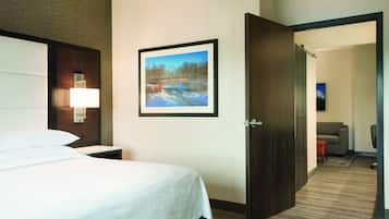 Una habitación de hotel con cama, mesita de noche, un cuadro en la pared y una puerta que da a otra habitación.