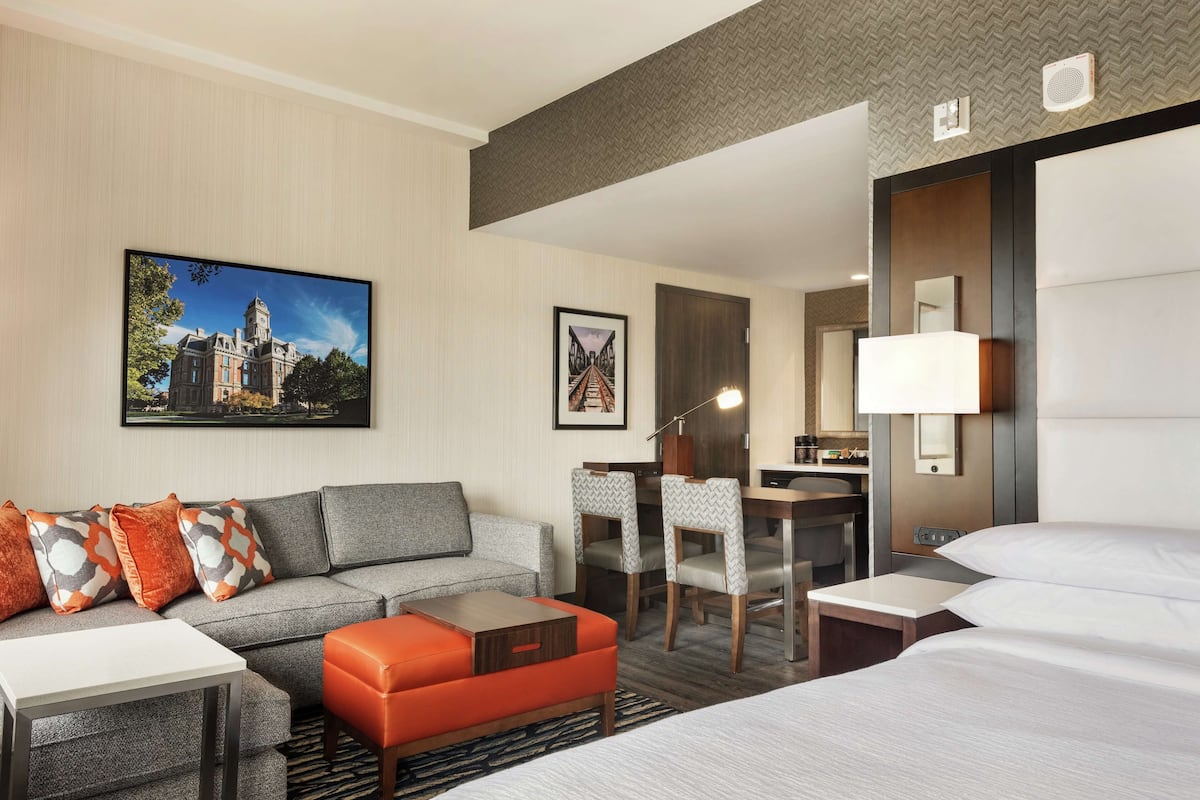 Une chambre d’hôtel avec un canapé gris, un pouf orange, un bureau avec des chaises, un lit et une grande fenêtre.