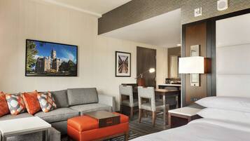 A hotel room with a grey sofa, orange ottoman, a desk with chairs, a bed, and a large window.