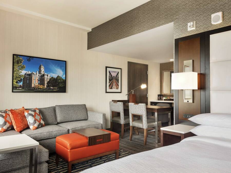 A hotel room with a grey sofa, orange ottoman, a desk with chairs, a bed, and a large window.