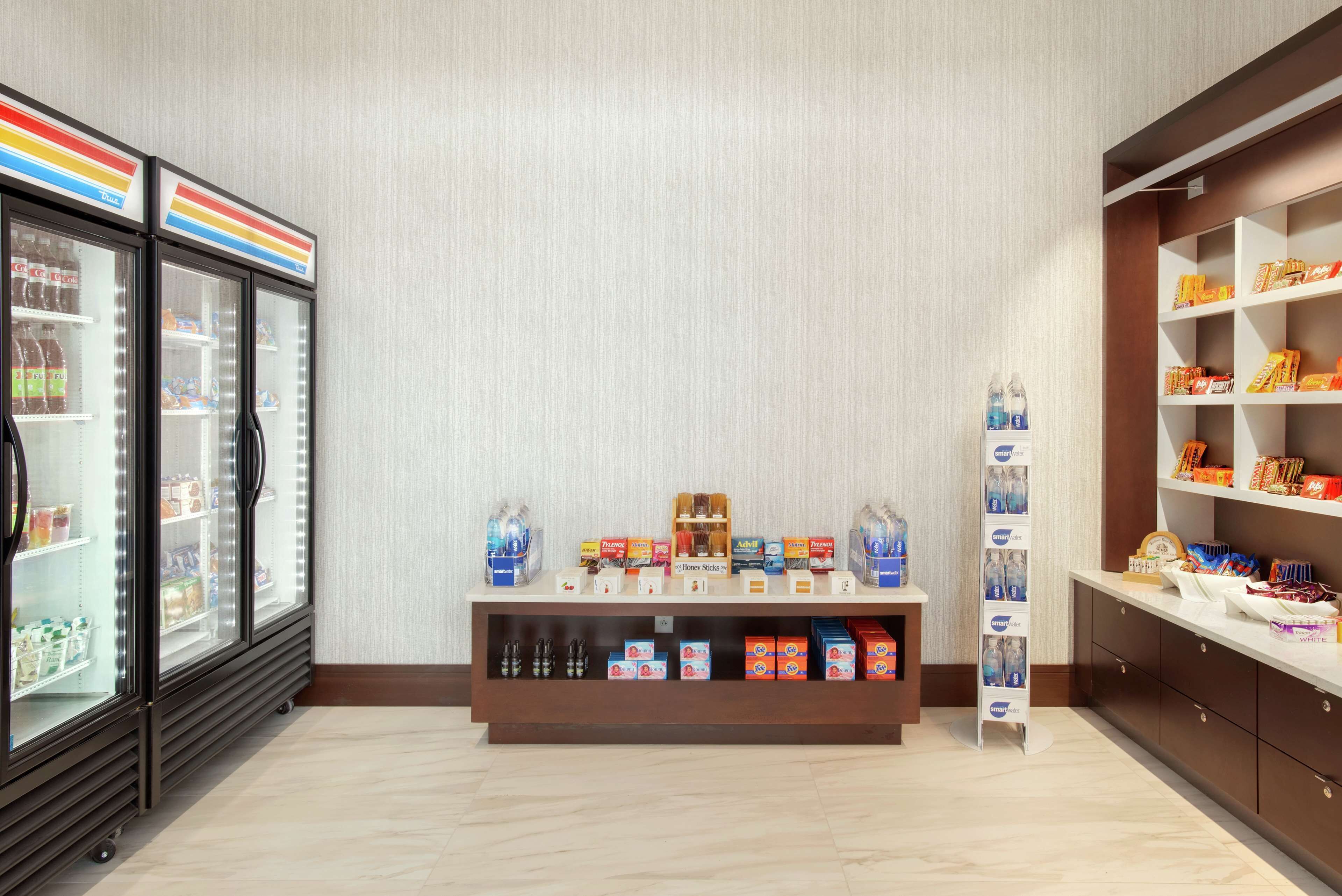 A hotel lobby with a refrigerated display case, a beverage dispenser, and a shelf with snacks and toiletries.