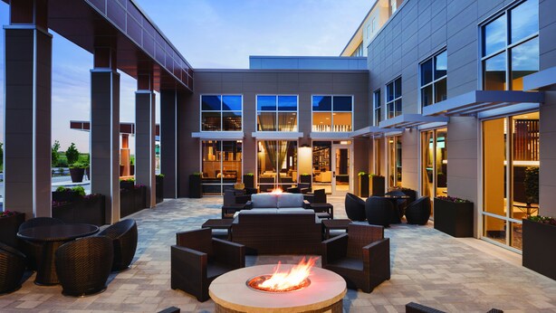 Un patio al aire libre con una fogata, mobiliario de mimbre y un edificio moderno al fondo.