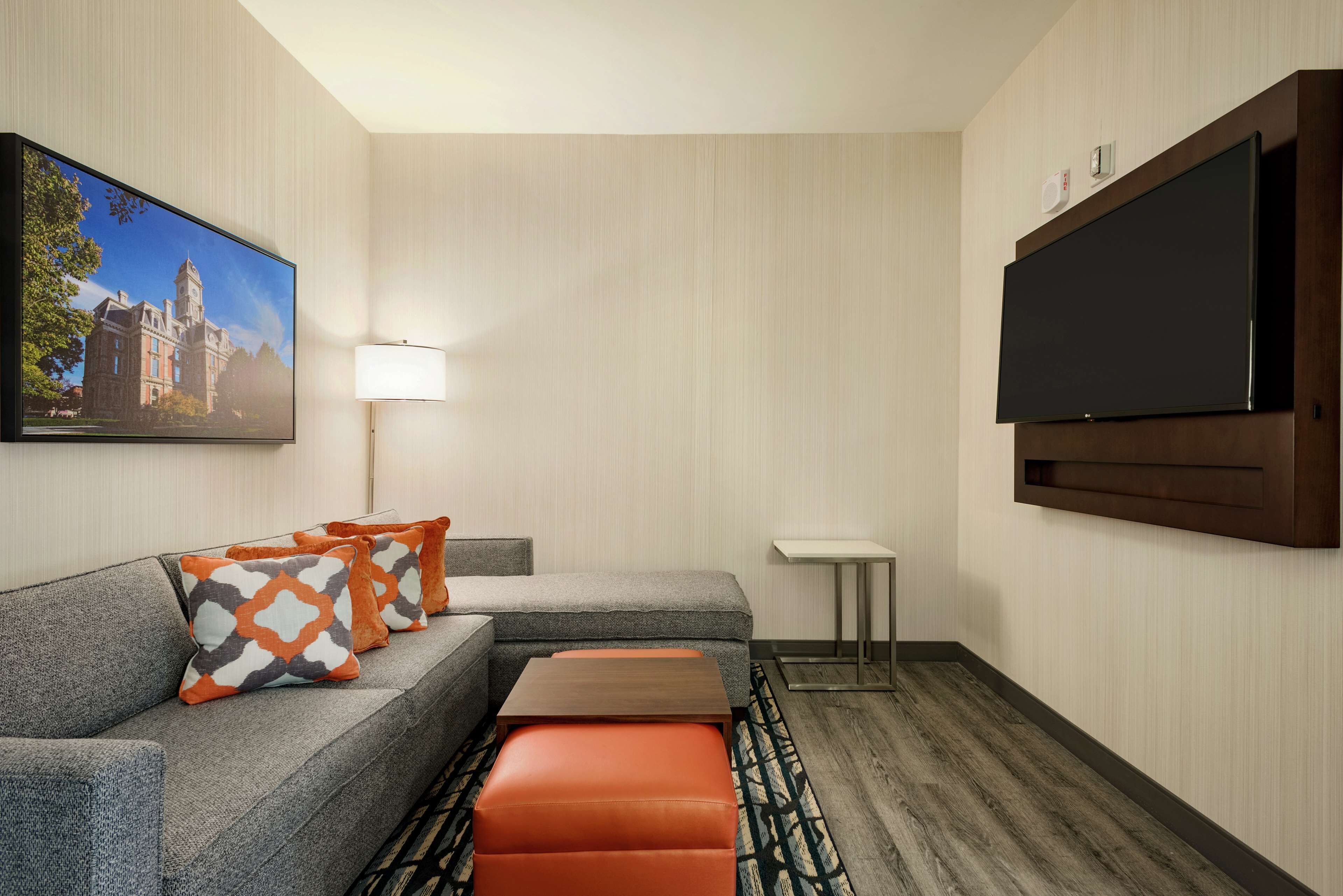 A hotel room with a grey sofa, orange and grey patterned pillows, a wooden coffee table, a flat-screen TV, a standing lamp, and a framed picture of a building.