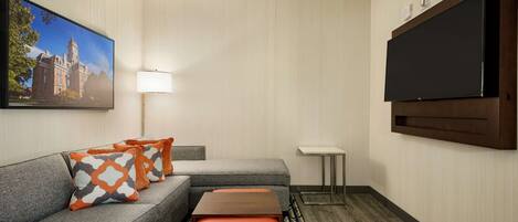 A hotel room with a grey sofa, orange and grey patterned pillows, a wooden coffee table, a flat-screen TV, a standing lamp, and a framed picture of a building.