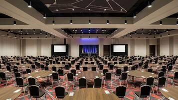 Une grande salle de conférence équipée de plusieurs tables et chaises, d’une scène avec un rideau bleu et d’un écran de projection.