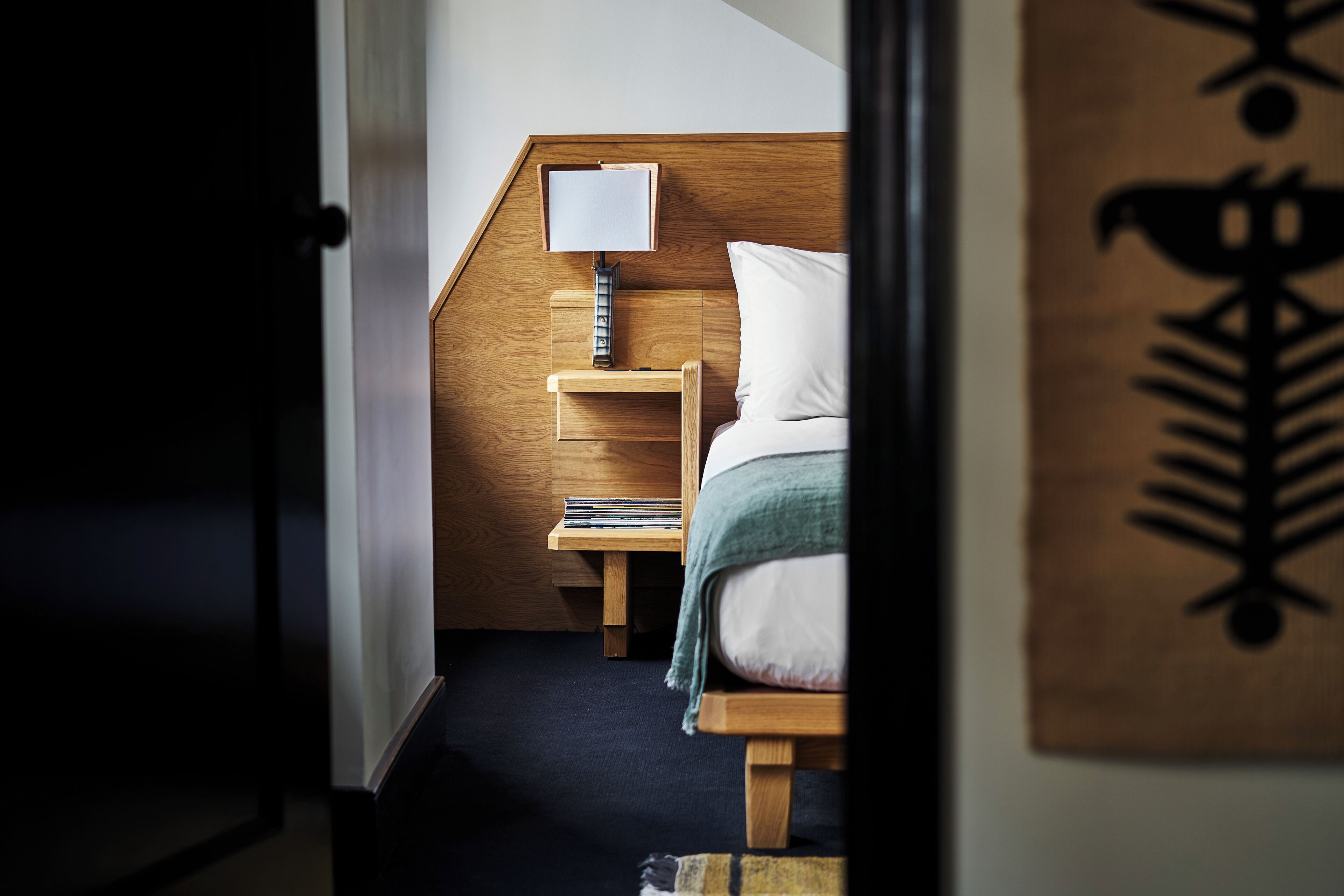 Un dormitorio con cabecera de madera, mesita de noche con lámpara, cama con ropa blanca y una pequeña estantería con libros.