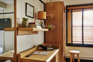 A wooden desk with a telephone, papers, and a remote control. There are two wooden shelves above the desk, one holding a lamp and a plant, and the other holding a basket with apples. A window with blinds is visible to the right.