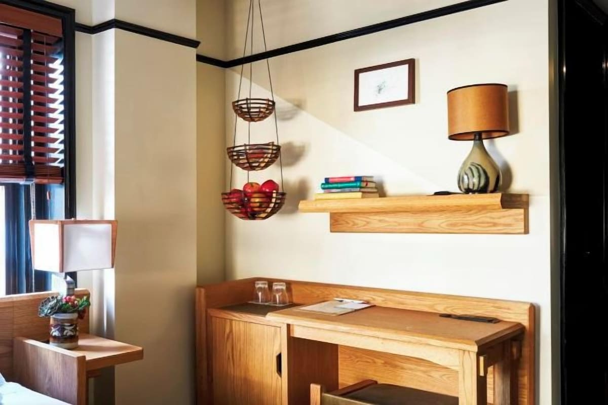 Una camera d'albergo con una scrivania in legno, una lampada, una libreria e una finestra con persiane.