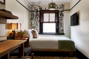 A bedroom with a bed, a desk with a lamp, a window with blinds, and a wall with a mural.