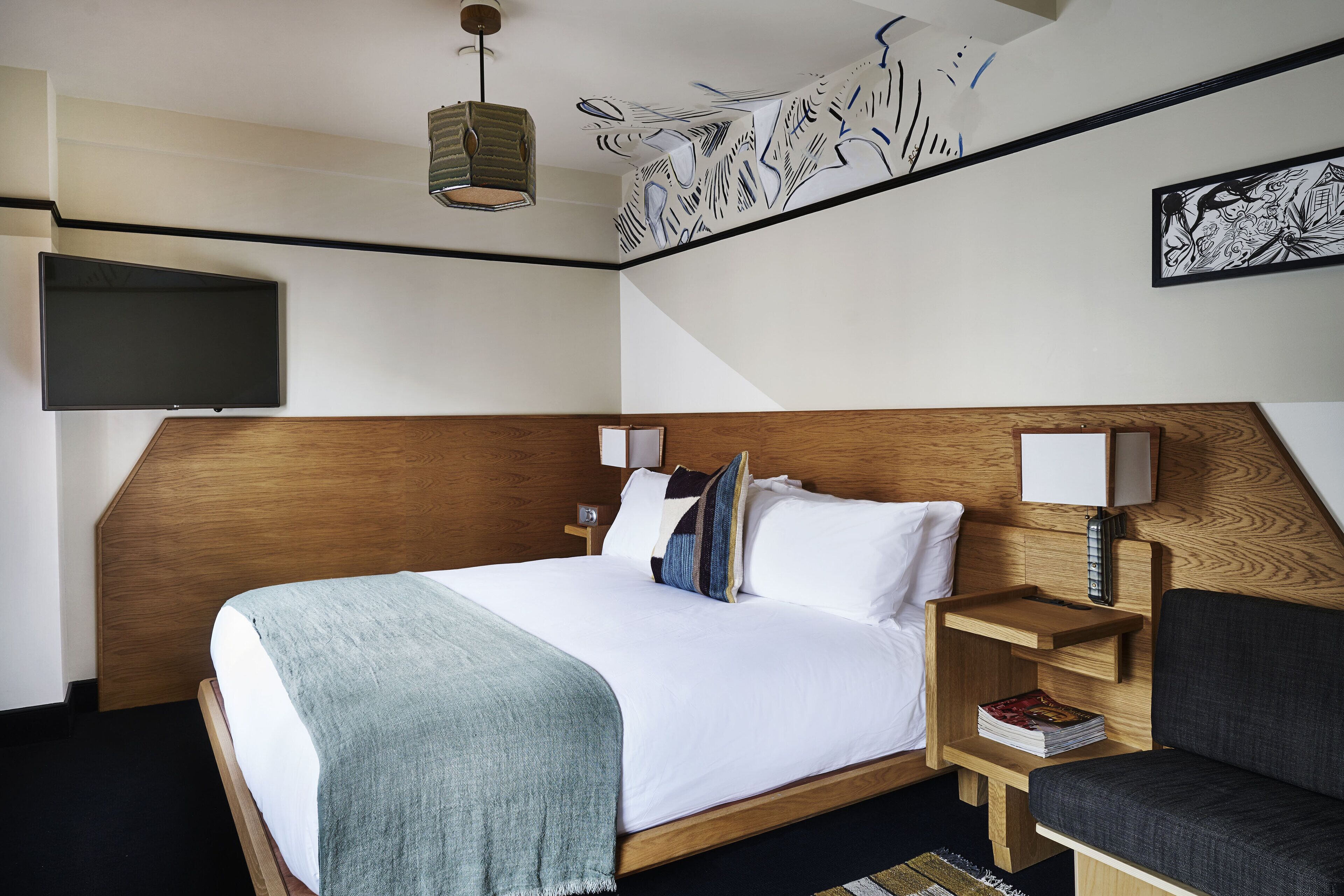 A hotel room with a wooden headboard, a bed with white linens, a grey throw blanket, a bedside table with a lamp, and a wall-mounted TV.