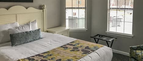 A neatly arranged bedroom with a bed, pillows, a bedside table, and two windows with blinds.