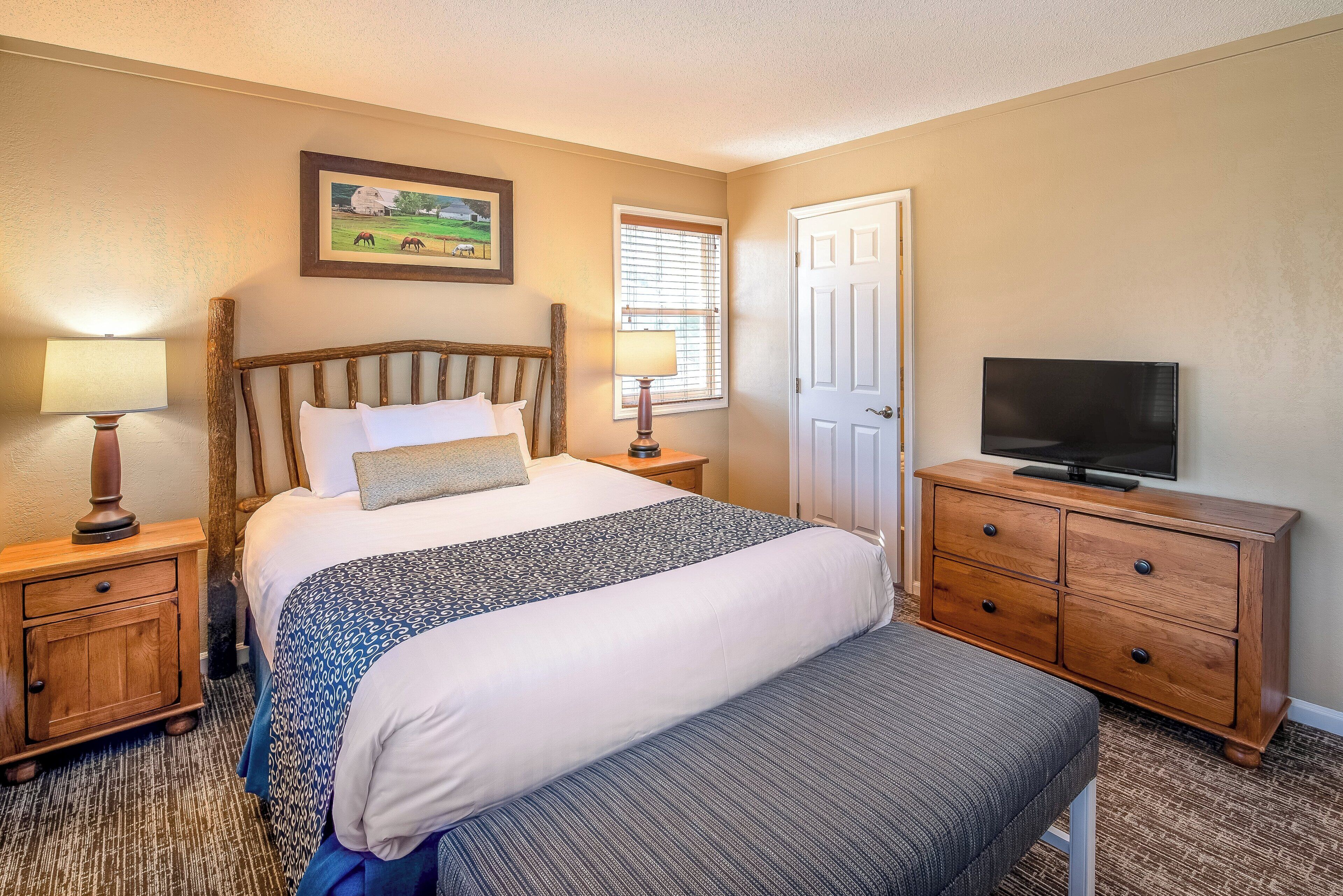 Une chambre d’hôtel avec un lit, une télévision, une commode en bois et une fenêtre avec des stores.