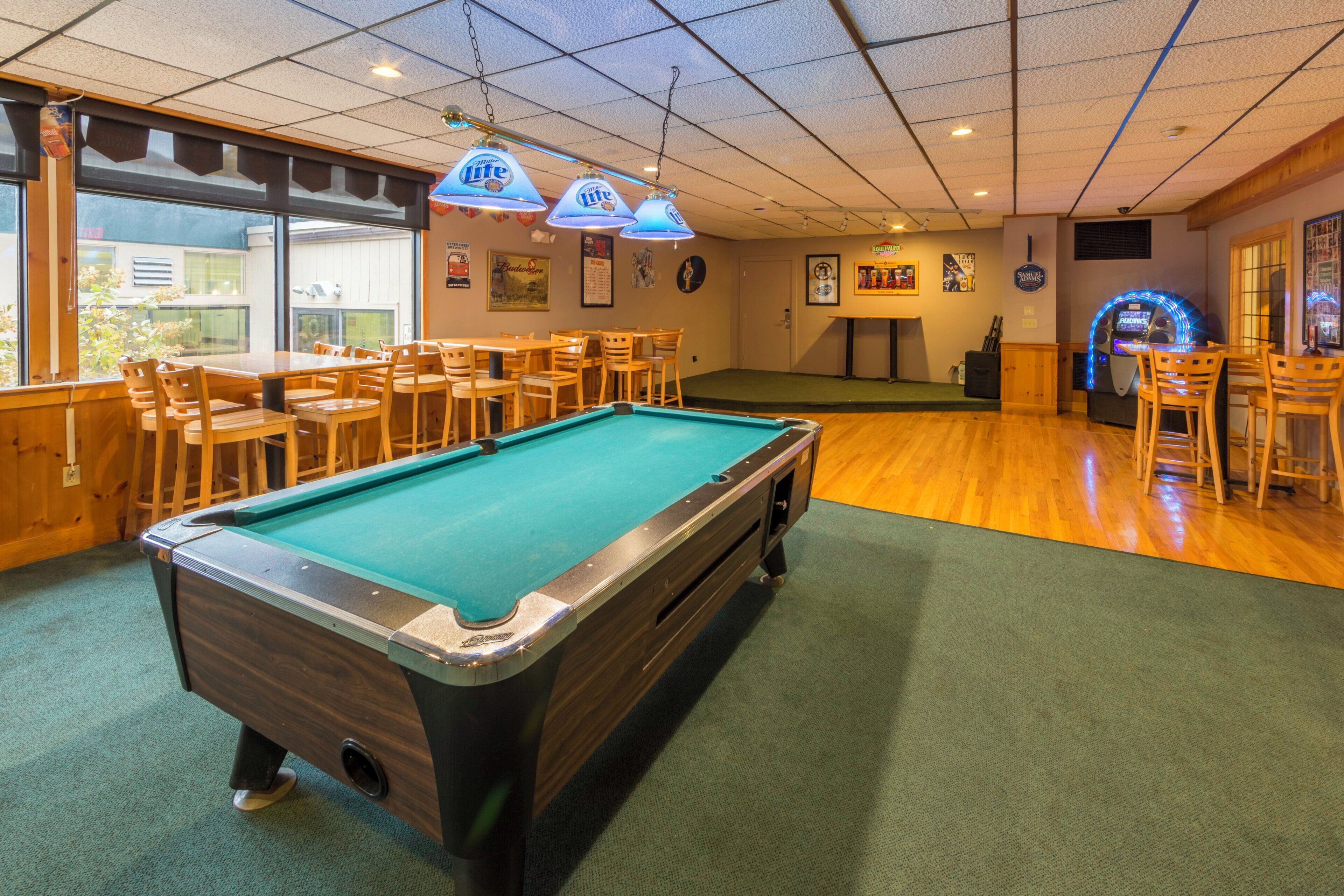 Mesa de billar en una sala con sillas y mesas de madera, y una zona de bar con taburetes.