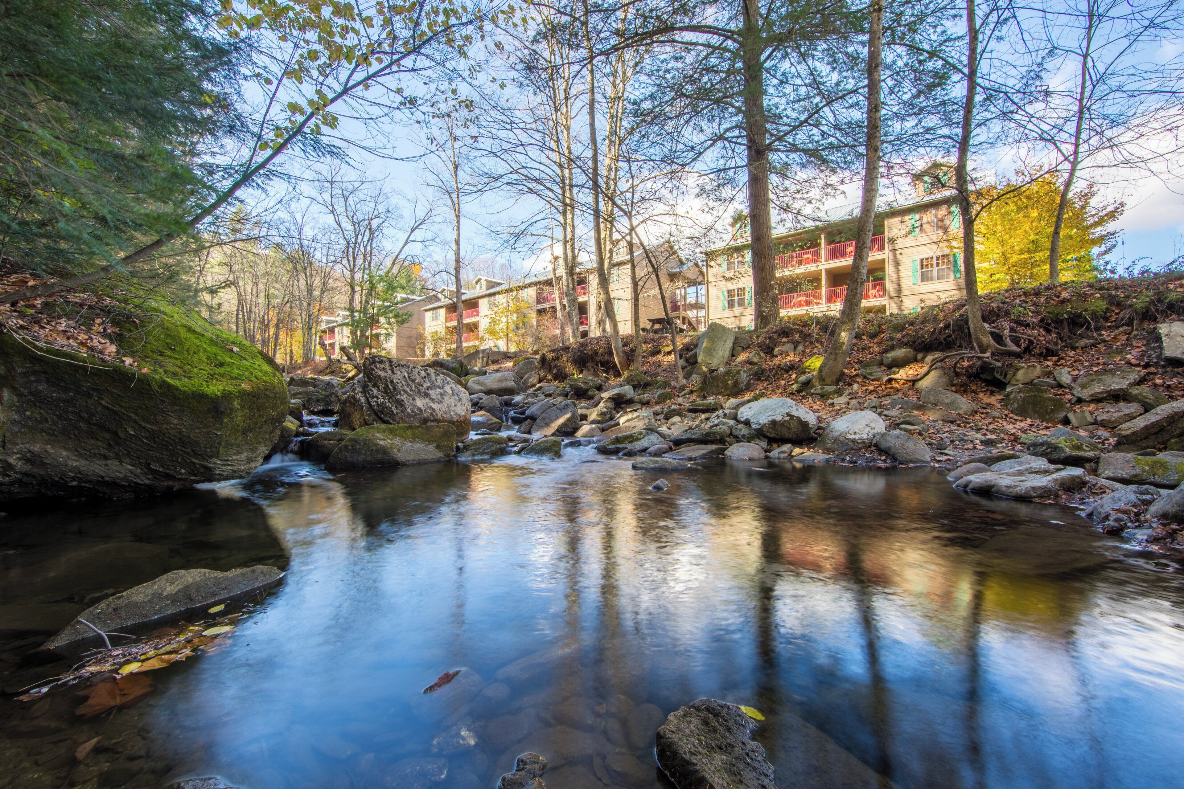 A tranquil stream with rocks and trees, a building with balconies and a red railing, and a clear sky.