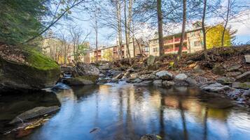 A tranquil stream with rocks and trees, a building with balconies and a red railing, and a clear sky.