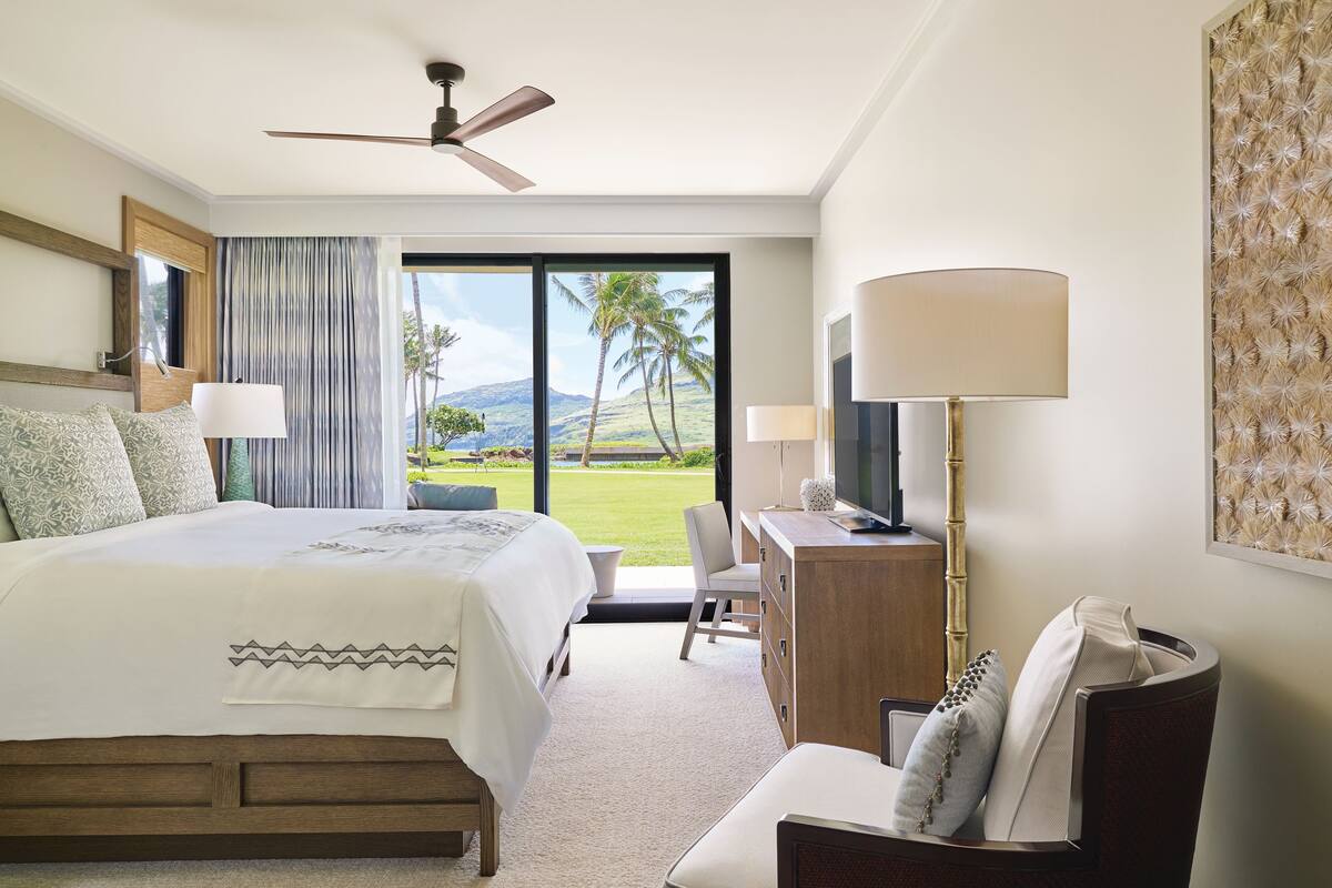 Une chambre d’hôtel avec un grand lit, un bureau avec une télévision, une chaise et une vue sur un paysage tropical par la fenêtre.