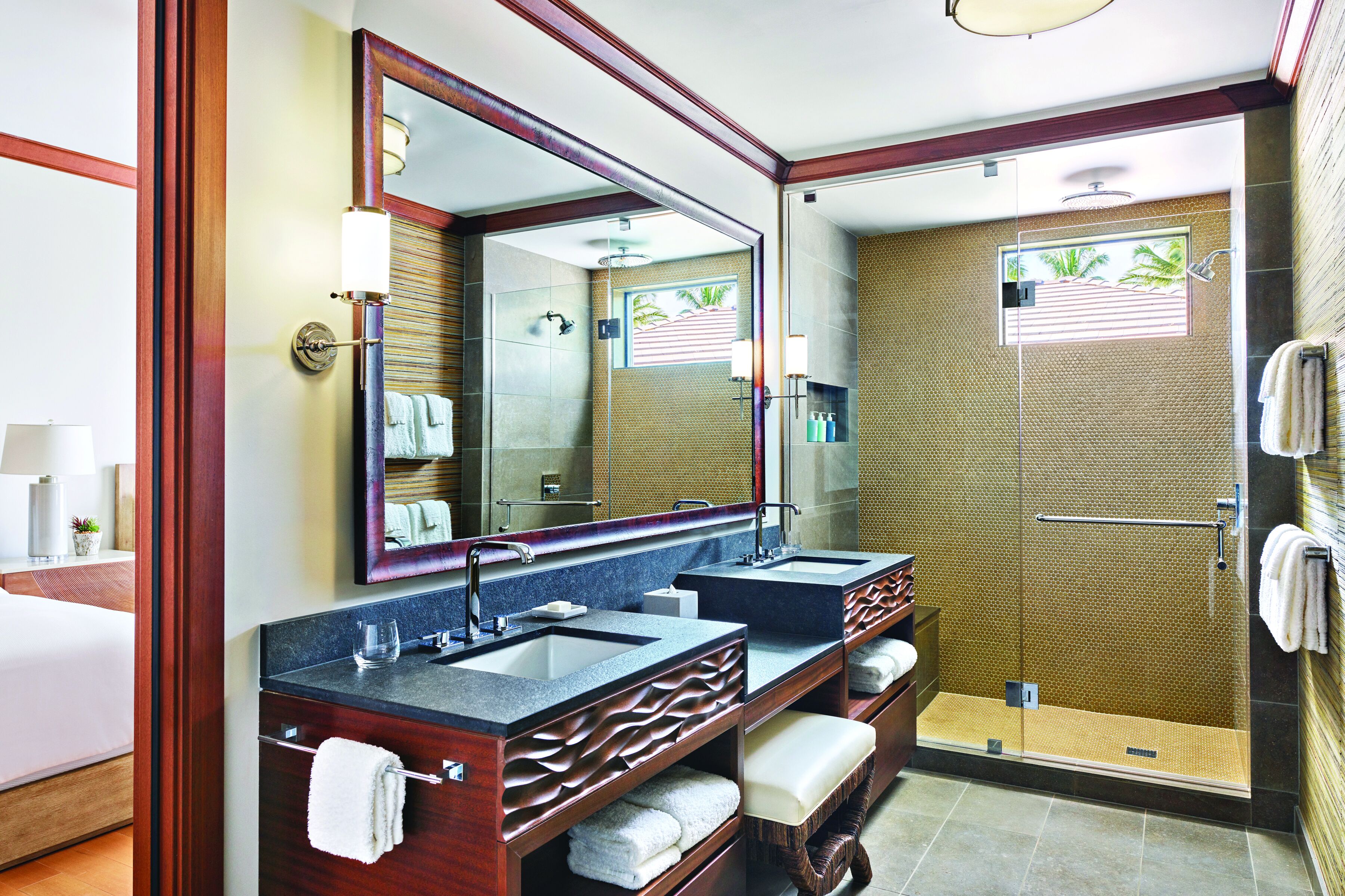 A hotel bathroom with a large mirror, two sinks, a shower, and a bed.