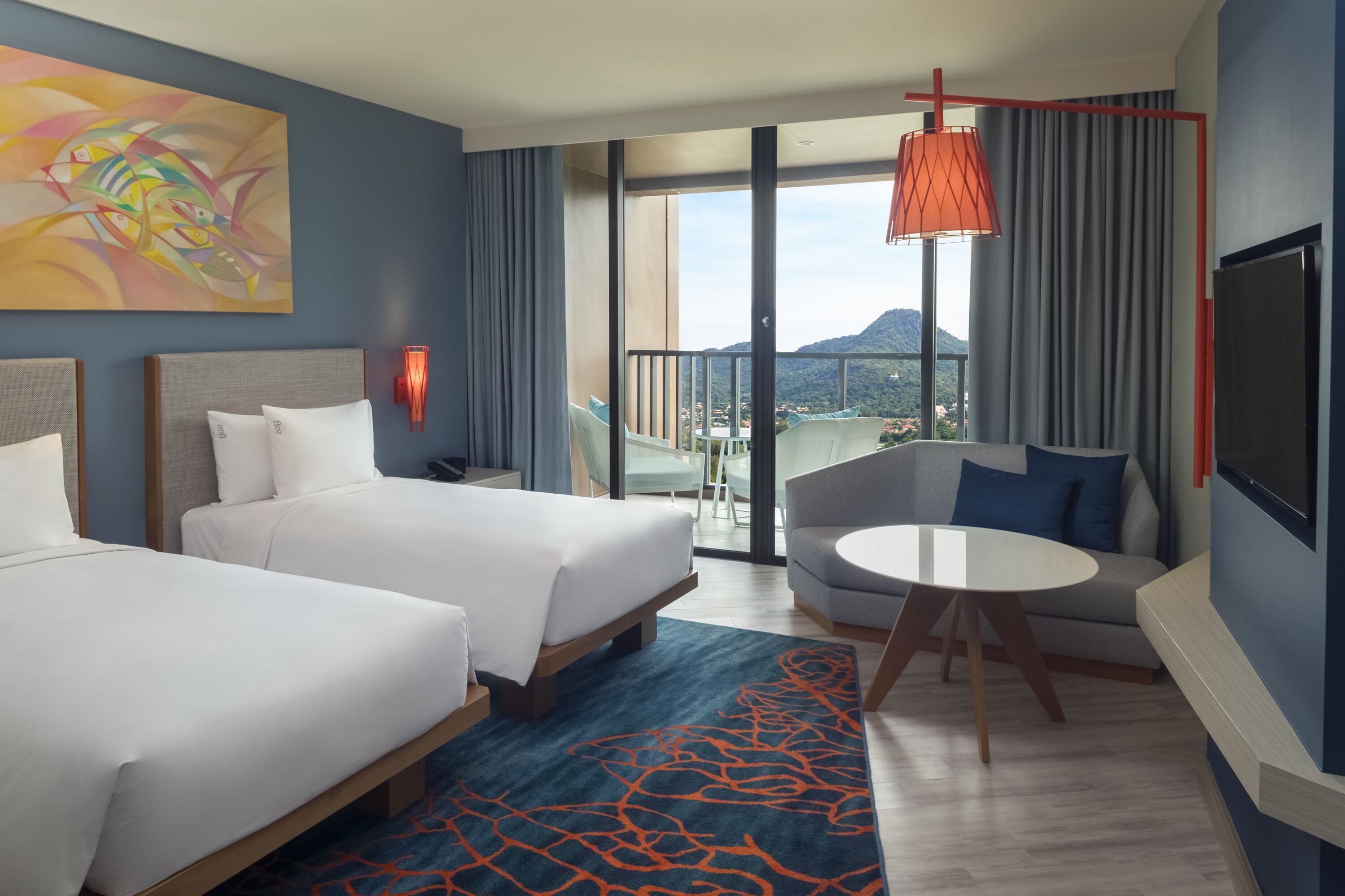 A hotel room with two beds, a sofa, a small table, a TV, and a balcony with a view of the mountains.