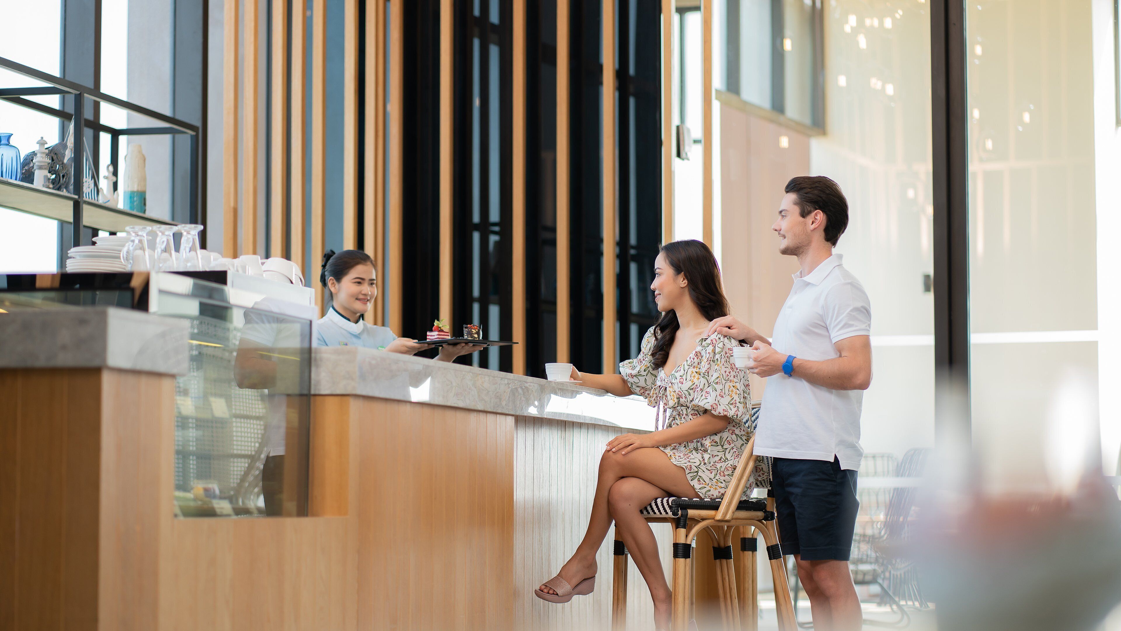 Un área de recepción moderna de hotel con un mostrador de madera, una mampara de vidrio y vistas al interior a través de grandes ventanales.