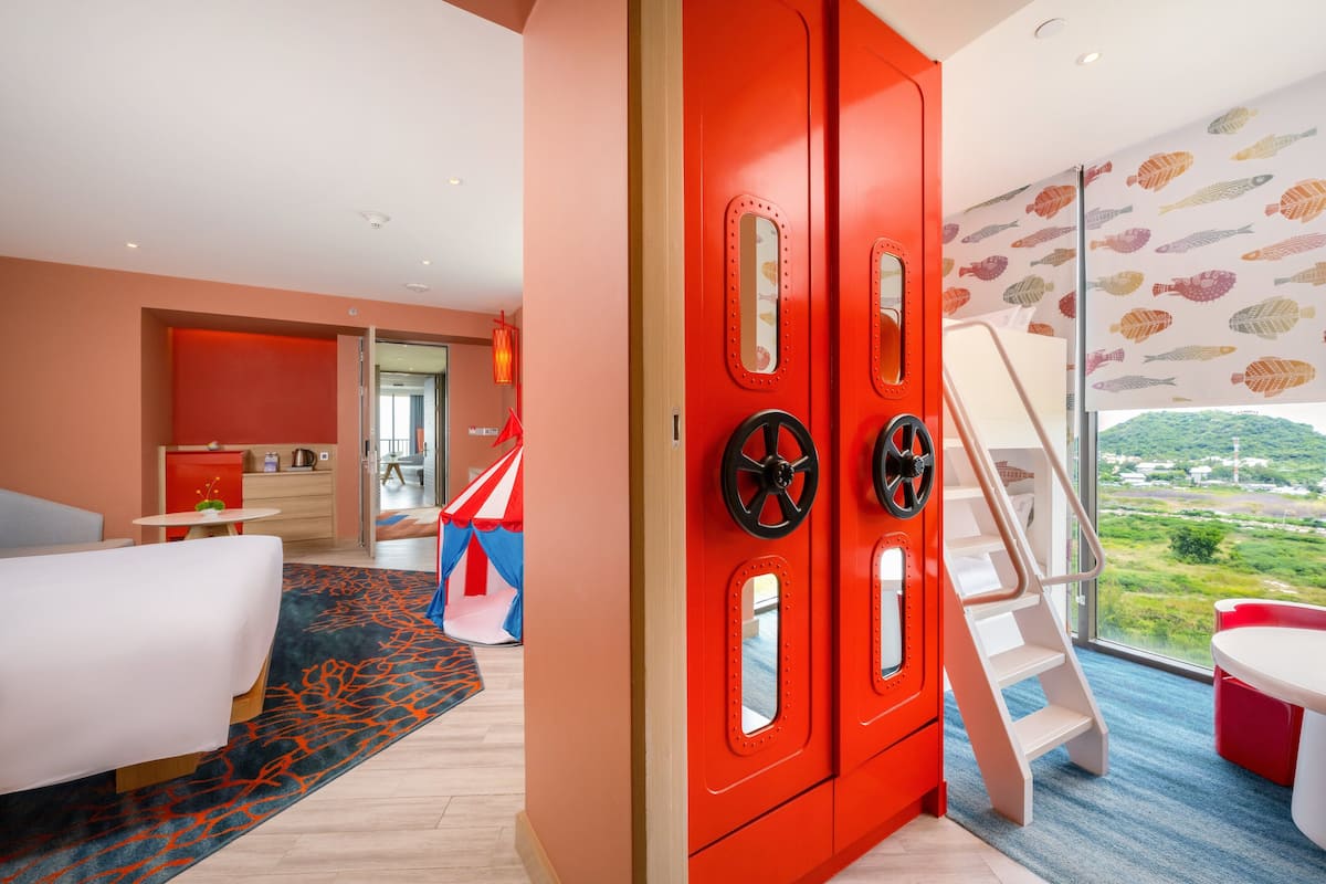 A modern hotel room with a red wardrobe, a bed, a desk, and a view of the countryside.