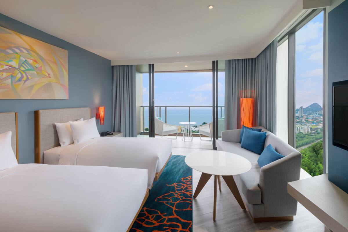 A hotel room with two beds, a sofa, a small table, and a balcony with a view.