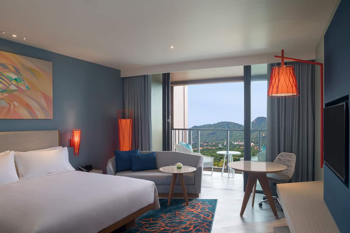 A hotel room with a bed, a sofa, a small table, and a balcony with a view of the city.