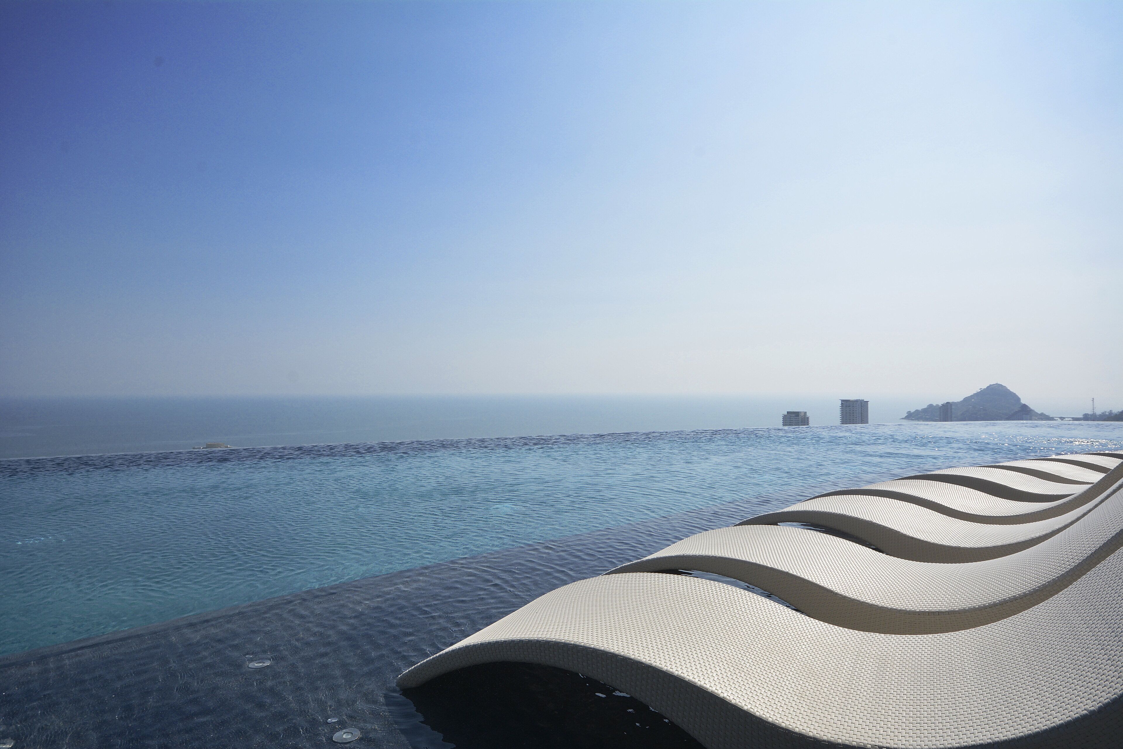 A poolside lounge with a view of the ocean and distant buildings.