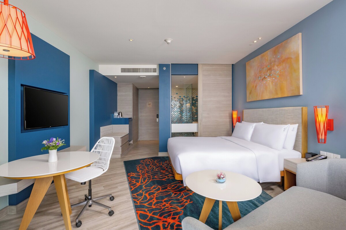Quarto de hotel com cama, escrivaninha, cadeira, televisão e um quadro na parede.