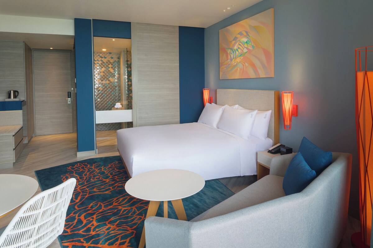 Quarto de hotel moderno com uma cama grande, um sofá, uma mesinha e uma área de estar separada.