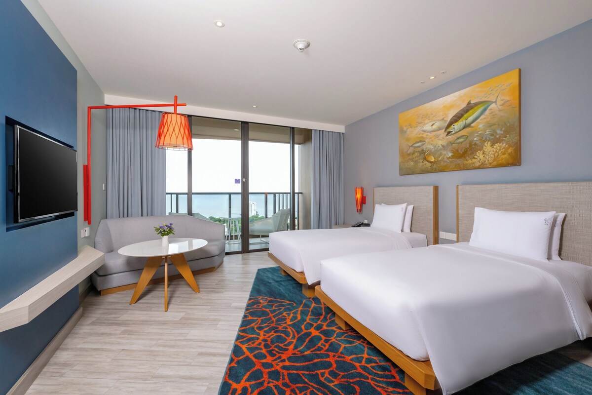 Quarto de hotel com duas camas, televisão, varanda com vista e um quadro na parede.