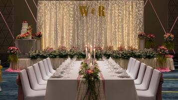 A long banquet table set for a wedding reception with a backdrop featuring 'W&R' and fairy lights.