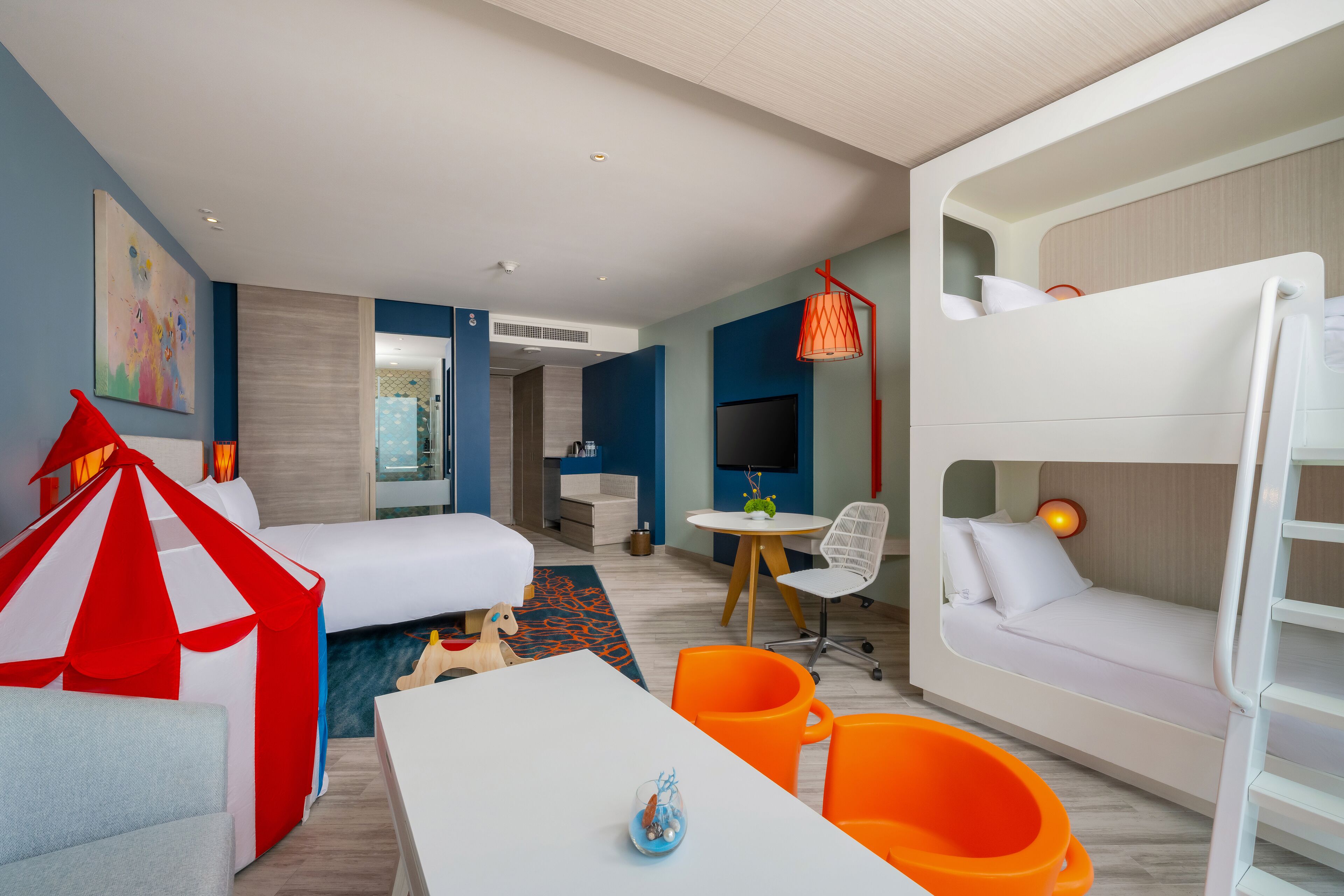 A modern hotel room with a bunk bed, a white sofa, a red and white tent-like structure, a round table, and orange chairs.