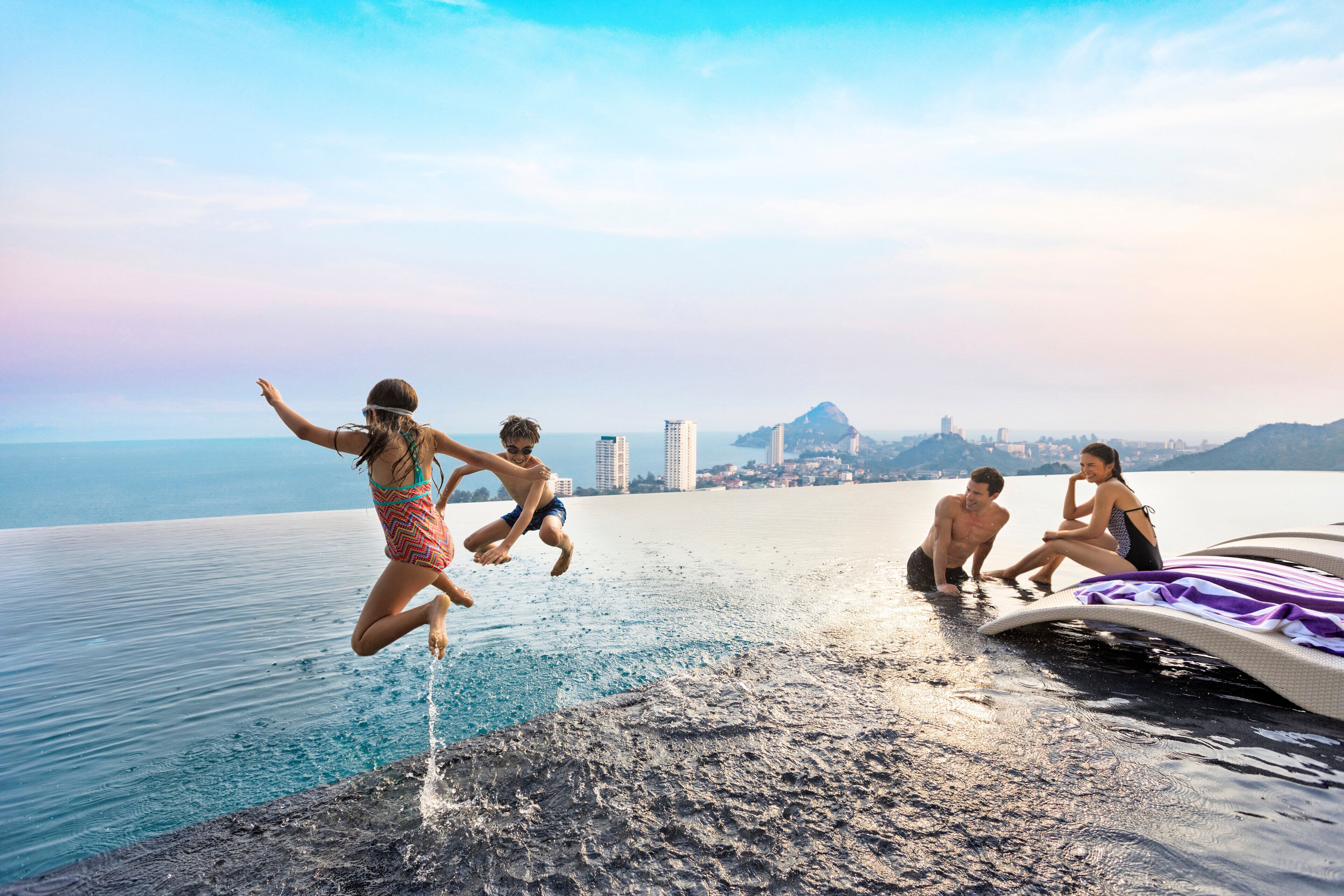 A group of people jumping into water from a high platform, with a cityscape and mountains in the background.