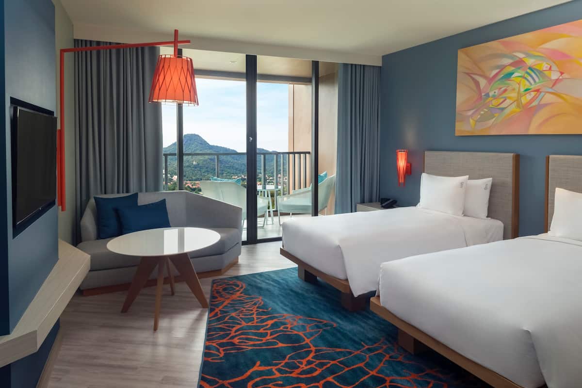 A hotel room with two beds, a sofa, a round table, and a balcony with a view of a mountain.