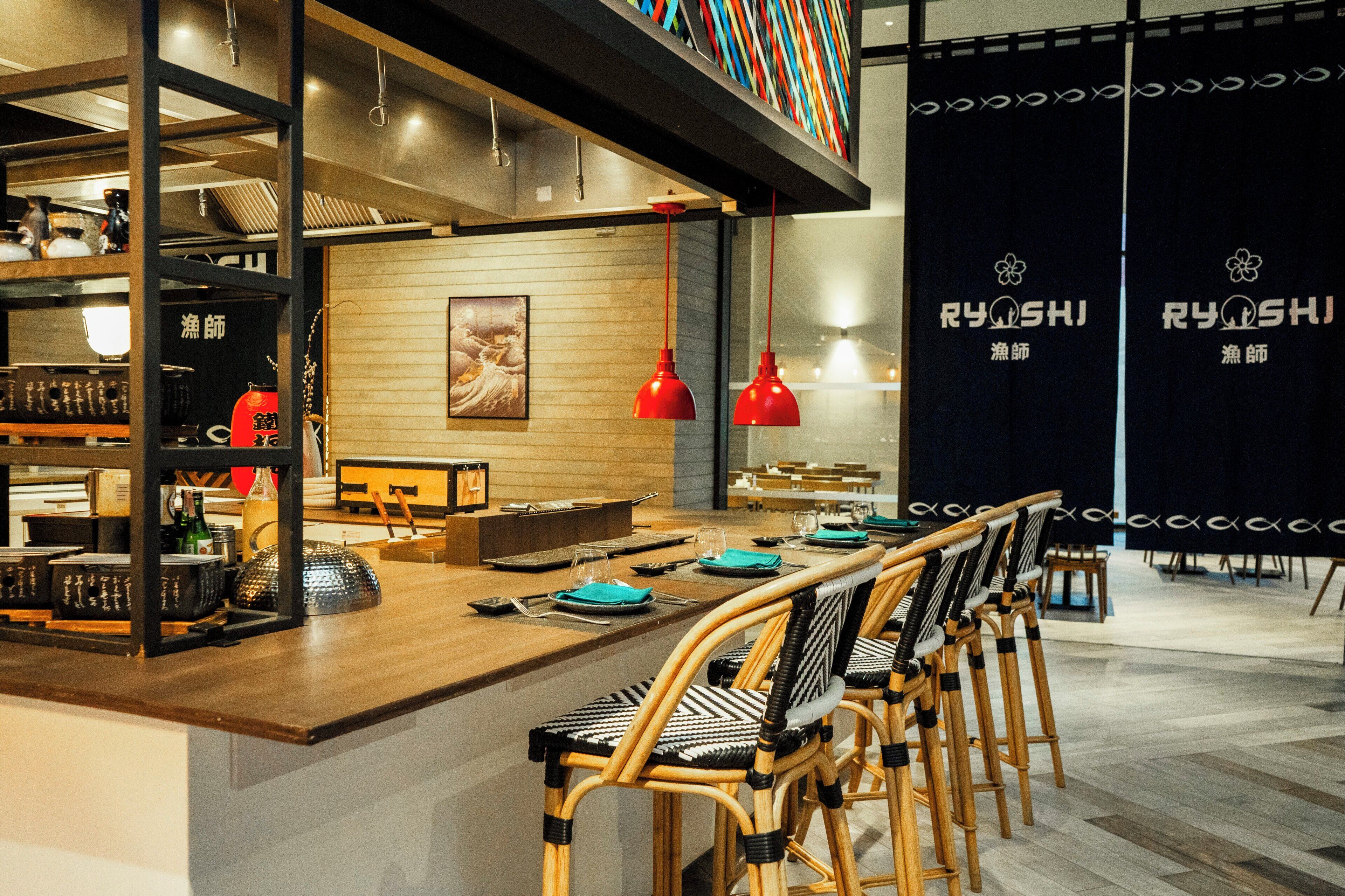 A restaurant interior with a wooden counter, red pendant lights, and bamboo chairs.
